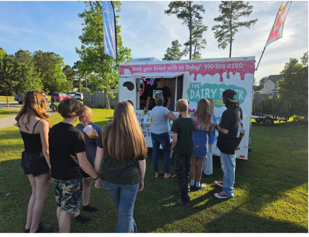 A line of kids and grown-ups waiting at The Dairy Duck's serving window on a grassy lawn at an outdoor event
