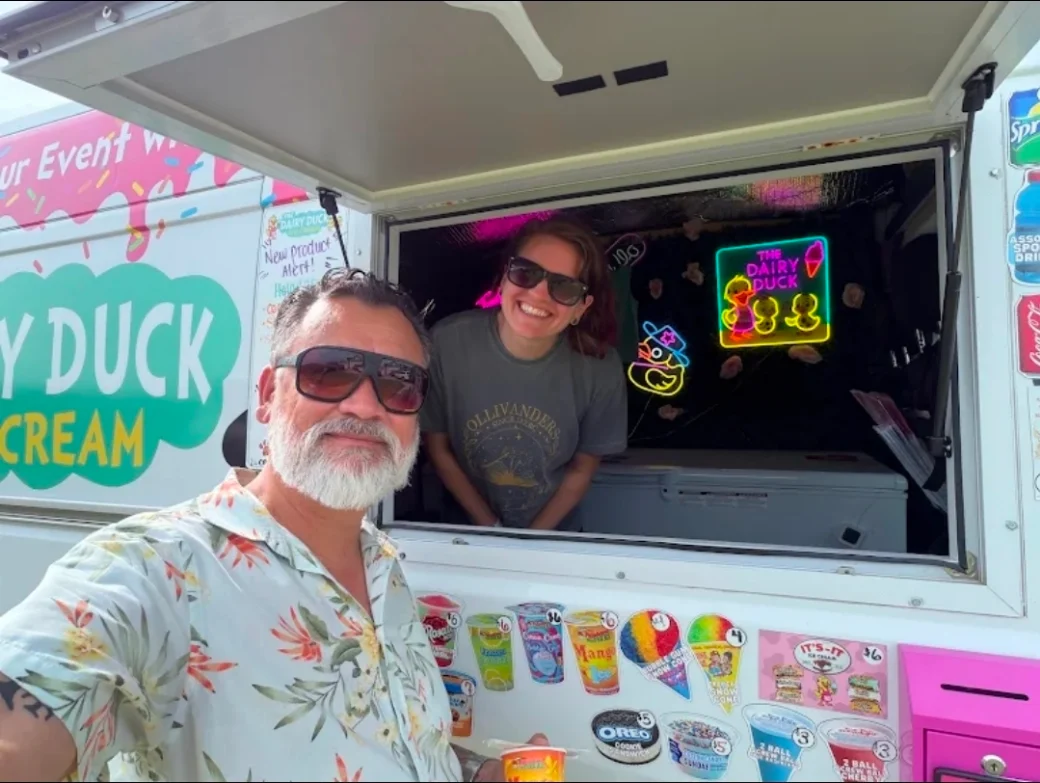 Two of the Dairy Duck crew at the serving window, one standing outside and one leaning out of the window, with the menu stickers along the side of the truck
