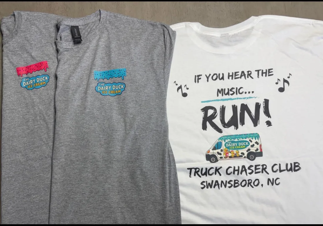 Three Truck Chaser Club shirts: two grey tees front side up, each with a small Dairy Duck Ice Cream logo on the chest, and a white tee turned over to show the back print reading If you hear the music, RUN, above the spotted Dairy Duck van and Truck Chaser Club Swansboro NC