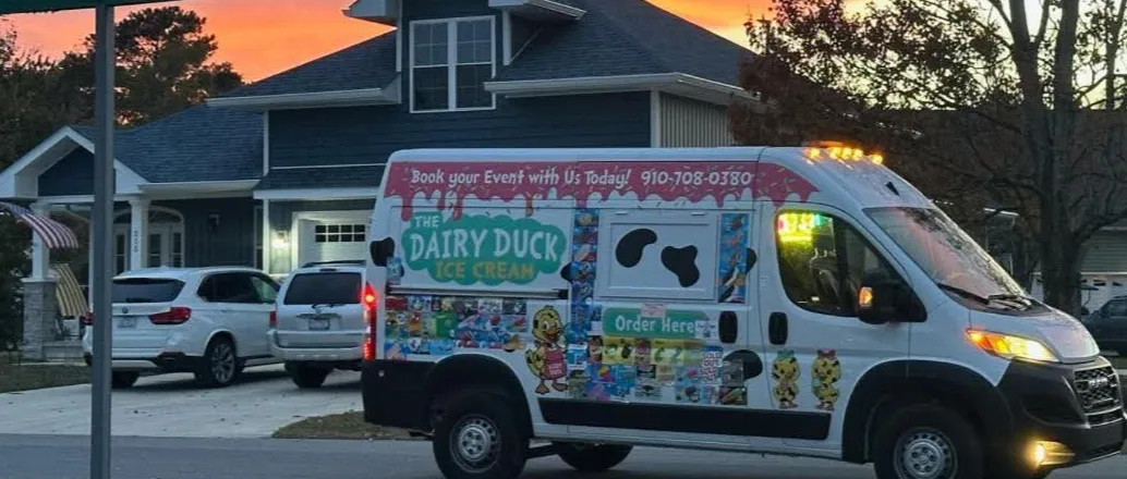 The Dairy Duck van parked on a neighborhood street at sunset beside a Duck Haven street sign, its Book your Event with Us Today banner and menu stickers lit up along the side