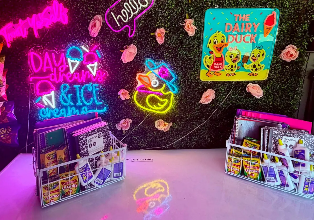 Inside the serving window: neon signs reading Day dreams and ice creams, hello, and a glowing yellow duck on a hedge wall, beside a carved Dairy Duck sign, with two wire baskets of crayons, glue and composition books collected for a school supply drive