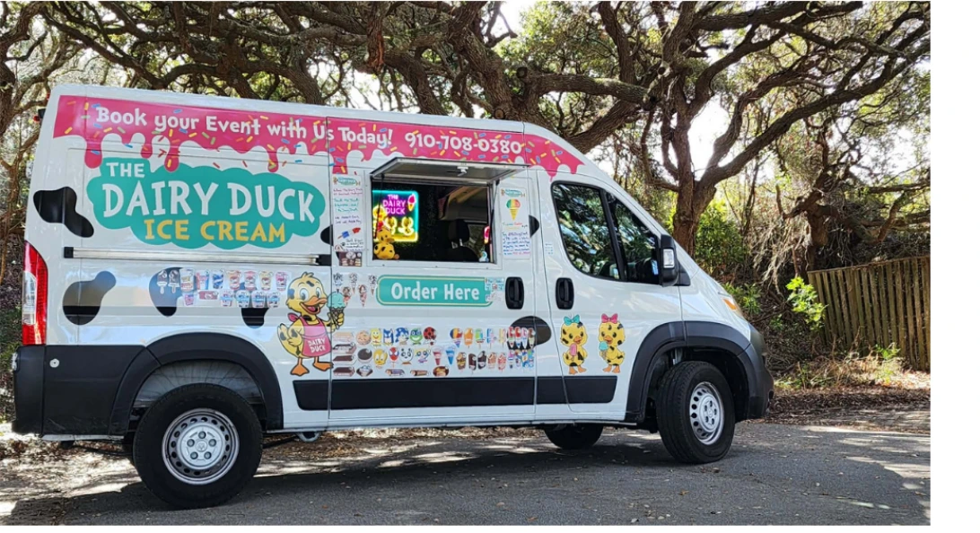 The Dairy Duck van parked under live oaks, its serving window open with an Order Here sign. A white van in black cow spots with a pink drip banner and the teal Dairy Duck Ice Cream logo